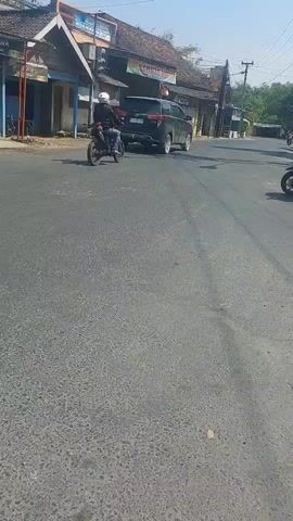 The video shows a street scene with several motorcycles and a car. The road is paved and appears to be in a semi-urban or rural area, with buildings and trees visible on the sides. The sky is clear and the lighting suggests it is daytime.

In the foreground, a motorcycle is moving from left to right, partially obscuring the view of the road ahead. The rider is not clearly visible.

Further down the road, several other motorcycles and a dark-colored SUV are present. On the left side of the road, there are buildings that appear to be shops or businesses. Traffic signals are visible.

On the right side of the road, a woman wearing a hijab and patterned leggings is riding a white scooter with an orange box attached to the back. She is looking forward.

Several other individuals on motorcycles are also present, moving in various directions, some of them wearing helmets and jackets. The general atmosphere suggests normal traffic activity.
