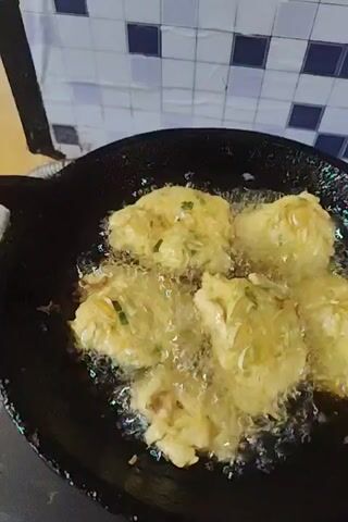 A close-up video shows several patties frying in hot oil in a black pan. The patties are irregularly shaped and light yellow-green in color, appearing to contain shredded vegetables. The oil is bubbling vigorously around the patties. A small section of a white and blue tiled wall is visible in the background. The pan is sitting on a stovetop.