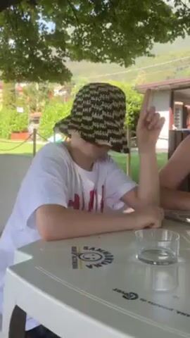 Two people are seated at a white plastic table outside. The person on the left wears a hat that covers their face and a white t-shirt with a red design. They are raising their index finger and moving their hands. The person on the right is wearing sunglasses and a blue tank top. There is a glass and a sippy cup on the table. Trees and a building are visible in the background.