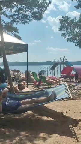 This is a daytime, vertical, medium-wide shot of a beach scene. In the foreground, two people lie in a hammock, viewed from the knees down. The hammock is striped blue and yellow and is suspended between trees. The beach is sandy with scattered tree roots and shadows cast from trees overhead. In the mid-ground, there are several beach chairs with people lying on them. Further back, there is a body of water, likely a lake, with people in the distance. Adjacent to the water is a large structure with stairs and a slide, and a pink, domed tent-like structure with tables and chairs underneath. A large tree trunk dominates the right side of the frame. The sky is blue with scattered white clouds.