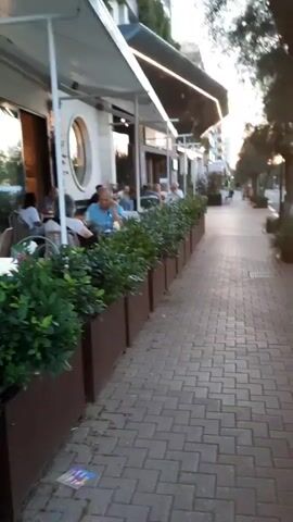 The video pans across an outdoor seating area of a restaurant on a paved sidewalk. Diners are seated at tables with white tablecloths, some eating and some talking. A man in a blue shirt is prominently visible, leaning on his hand. The restaurant has a white facade with a large circular window and awnings providing shade. Planters filled with dense green bushes line the edge of the seating area, separating it from the sidewalk. The street continues to the right, with trees and buildings visible in the background. The lighting suggests it is late afternoon or early evening.
