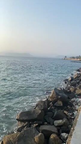 The video is a close-up shot of the ocean from a rocky shore. The water is a deep blue-green with small waves lapping against the rocks. The sky is a pale, hazy blue, with the sun visible in the distance, casting a shimmering reflection on the water. In the background, distant landforms can be seen through the haze. The scene is calm and serene, with no people or objects other than the rocks and the water.