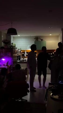 A dimly lit indoor scene, possibly a kitchen and living area, with several individuals present.  The lighting is predominantly purple and yellow from string lights. In the foreground, a person sits in a dark chair, facing away from the camera. To the left, another person is partially visible, also seated. In the mid-ground, two men stand near what appears to be a kitchen counter, with one man in a light-colored outfit and the other in dark clothing. Behind them, in the kitchen area, cabinets and tiled backsplash are visible. To the far right, a third person is partially visible standing on what might be a staircase or elevated platform. The overall atmosphere suggests a casual gathering.
