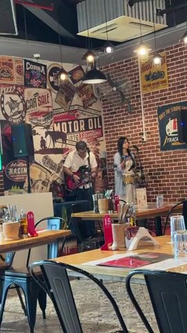 A man plays an electric guitar and sings into a microphone while a woman plays a drum and sings into a microphone in a restaurant. The restaurant has brick walls decorated with vintage posters and a metal ceiling. Tables with chairs are set up for diners. A sound speaker is on the left side of the frame.