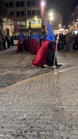 At night, a group of people dressed in blue pointed hats and red robes stand in a town square paved with cobblestones. They are surrounding two banners. The crowd is to their left and behind them.  The lights of the town illuminate the scene. One person is walking away from the group to the right of the frame. There are other people milling around in the background. A drumbeat can be heard.