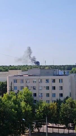 A video captures a plume of black smoke rising into a clear blue sky from behind a line of trees. In the foreground, a light-colored, multi-story building with many windows is visible. Lush green trees are in front of and around the building. The smoke appears to be emanating from an area beyond the trees and the building, suggesting a distant event. The overall scene is an outdoor urban or suburban environment on a sunny day.