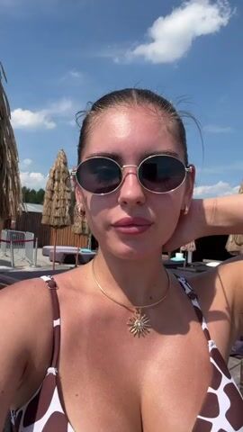 A young woman with dark hair pulled back in a bun, wearing sunglasses and a giraffe print bikini, poses for a selfie. She is outdoors in a lounge area, possibly a resort or beach club. In the background, there are lounge chairs, umbrellas, and cabanas. The sky is blue with some clouds, and the sun is shining brightly. The woman is looking at the camera and pouting. She is wearing a gold necklace with a sun pendant. Her skin is tanned, suggesting she has been in the sun. She adjusts her hair and then turns her head to the side, looking off-camera. The video appears to be a short, casual recording of the woman enjoying her time in the sun.
