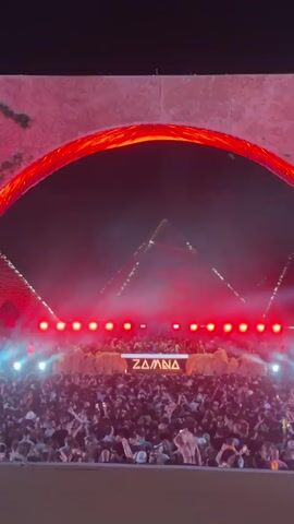The video shows an outdoor concert scene with a large crowd facing a stage. Behind the stage, the Egyptian pyramids are visible, illuminated in red light. The stage is marked with the word "ZAMNA."  Above the stage is a large, glowing red arch that resembles fire.  Bright spotlights shine out from the stage, adding to the vivid spectacle.  The crowd appears to be enjoying the performance.