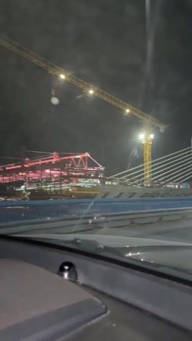 A night view from inside a vehicle shows a bridge under construction. A large yellow crane is visible, along with the cable-stayed structure of the bridge. The scene is illuminated by artificial lights.