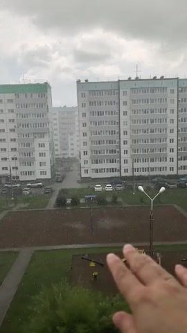 A person's hand is extended into the frame, reaching towards the viewer from the right side. The hand is slightly blurred, with a ring visible on the ring finger. In the background, tall apartment buildings are visible under a cloudy sky. It appears to be raining, with visible streaks of precipitation against the windows and falling in the courtyard between the buildings. There is a playground area in the foreground with some playground equipment. Cars are parked in the courtyard.