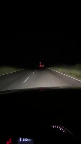 The video captures a first-person perspective from inside a vehicle driving on a dark, rural road at night. The headlights illuminate the road ahead, revealing a center white line and grassy verges on either side. In the distance, the taillights of another vehicle are visible, appearing red and slightly blurred due to the darkness and distance. The dashboard of the vehicle in which the video is being recorded is partially visible in the foreground, showing what appears to be a speedometer with markings up to 200 and a red needle indicating a speed of approximately 40. The sky is completely black, indicating it is nighttime. The overall environment is dark and rural, with no other visible structures or signs of civilization besides the road and the distant vehicle.