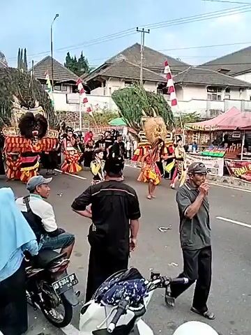 A group of people, some in elaborate costumes resembling mythical creatures and others in traditional attire, are participating in a parade on a street. The costumes are large and colorful, featuring masks with fierce expressions and headdresses made of foliage. Some individuals are carrying flags. Spectators are gathered on the side of the road, watching the procession. In the background, there are buildings with tiled roofs and trees. The sky is overcast. The scene appears to be a cultural or festive event.