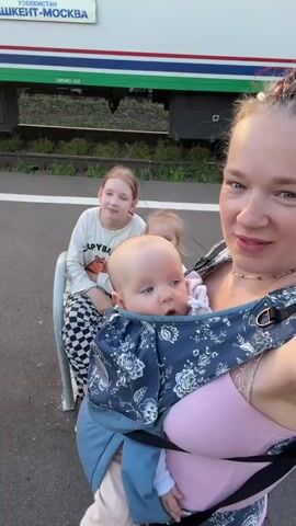 A woman with braided hair and a pink top holds a baby in a blue floral baby carrier. She is taking a selfie with two other children in the background. A younger child with blonde hair is sitting next to a teenage girl with blonde hair. The teenage girl is wearing a white long-sleeved shirt and checkered pants. The younger child is wearing a light-colored outfit. They are all sitting on a bench next to a train. The train is white with green and blue stripes. The background shows train tracks and greenery. The lighting suggests it is daytime.