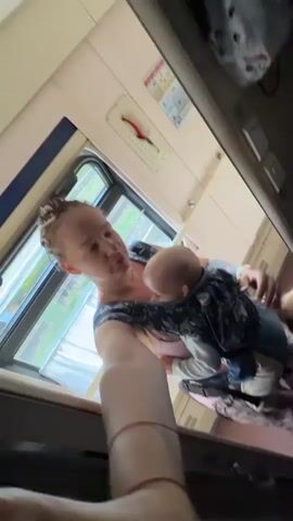 A woman with her hair pulled back is holding a baby in a carrier. She is wearing a dark-colored top with a floral pattern. The baby is wearing a light-colored outfit. They are inside a train car, with a window behind them showing greenery outside. The woman is holding up a phone to take a selfie. There is another person, partially visible in the background, also appearing to be on the train.