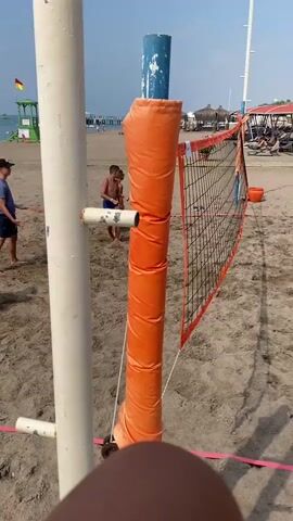 A video taken at a beach shows two boys playing volleyball near a net. The camera is focused on a volleyball net and pole, with two boys in the background. One boy is wearing a blue shirt and shorts, and the other is wearing swim trunks. Both are facing away from the camera. The beach has sand and some vegetation. The sea is visible in the distance, as is a lifeguard tower. The sky is blue and clear.