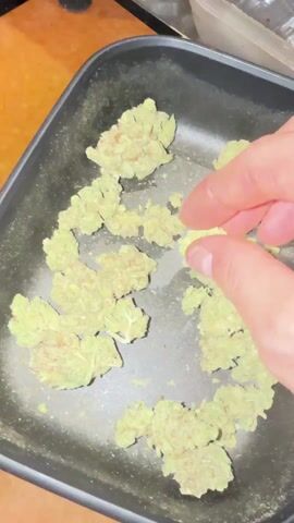 A hand picks up and examines a light green bud of cannabis flower. The bud is dense with visible trichomes. The flower is held above a small black tray containing several other buds of similar appearance. The background is blurry, but shows an orange surface.