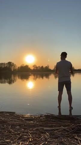 Here is a description of the video:
The video shows a person standing on the edge of a body of water at sunset. The person is silhouetted against the bright orange sun, which is reflected in the water. The person is wearing shorts, a light-colored tank top, and a baseball cap. The shoreline is covered in dry reeds and other vegetation. Later in the video, a child walks up and pokes the water with a stick.