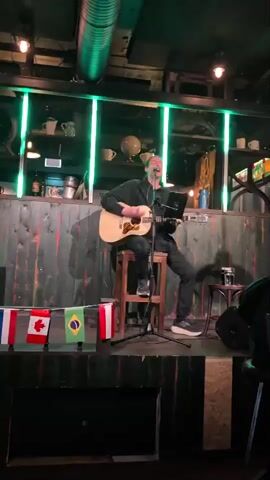 A man sits on a wooden stool playing an acoustic guitar and singing into a microphone. He is wearing a black hoodie and black pants. He is on a stage decorated with flags from Canada and Brazil. Behind him is a wooden wall with green lights.