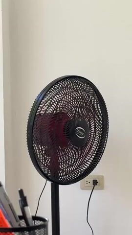 A black oscillating fan is shown from the front. The fan is plugged into a wall outlet, and the blades are spinning. The fan is on a stand, and its base is partially visible. In the foreground, a black mesh cup filled with pens and markers is visible. The background is a plain white wall. The video is shot from a medium distance, and the lighting is bright and even.