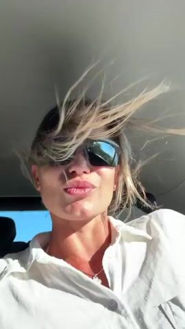 This is a close-up, first-person perspective video of a person inside a vehicle, likely a car. The subject is a woman with short, blonde hair that is being blown around by the wind, creating a dynamic effect. She is wearing dark sunglasses that reflect the sky and the surrounding environment, which appears to be sunny. Her lips are pursed, suggesting a playful or perhaps slightly exaggerated expression. She is wearing a white collared shirt.

The activity taking place is a ride in a vehicle, with the open window allowing the wind to blow through the car. The time of day is clearly daytime, indicated by the bright sunlight and the clear blue sky visible through the sunglasses' reflection. The weather appears to be warm and sunny, conducive to having the windows down.

While no specific text is visible in the frame, the location context provided is Rudinice, Montenegro. This suggests the scene is set in or around this area. The overall mood conveyed is one of freedom, fun, and enjoying the outdoors. The wind in her hair and the sunglasses evoke a sense of movement and carefree enjoyment, possibly during a road trip or a scenic drive. The angle of the shot and the focus on the subject's face and hair draw attention to her animated reaction to the wind.