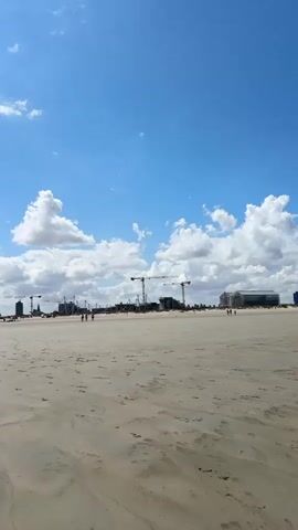 A wide, sandy beach stretches towards the ocean under a bright blue sky dotted with white clouds. In the distance, a construction site is visible, with several large cranes and building structures. A few people are scattered across the beach, some walking, others appearing to stand and observe. The waves gently lap at the shore.