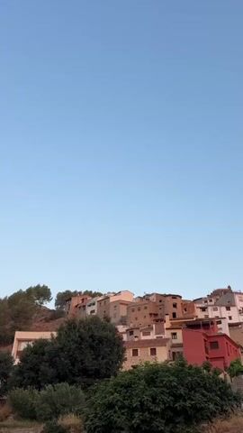 The video pans across a hillside with a small village built upon it. The sky is clear and blue, suggesting it is daytime. The village appears to be made up of older buildings, with some showing signs of age. Green trees and vegetation are scattered throughout the scene, with some growing in front of and around the buildings. In the distance, a rocky landscape can be seen, with the sun catching its reddish hues. The camera movement is slow and smooth, giving a sense of calm observation. There are no people visible in the video.