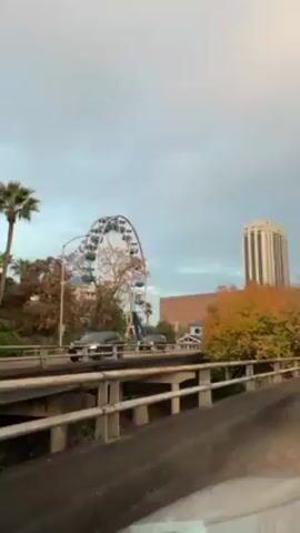 The video shows a view of a cityscape from a car driving on a bridge. There are several tall buildings in the background, including a Ferris wheel. The trees in the foreground have autumn colors. The sky is overcast. Cars are moving in both directions on the bridge.