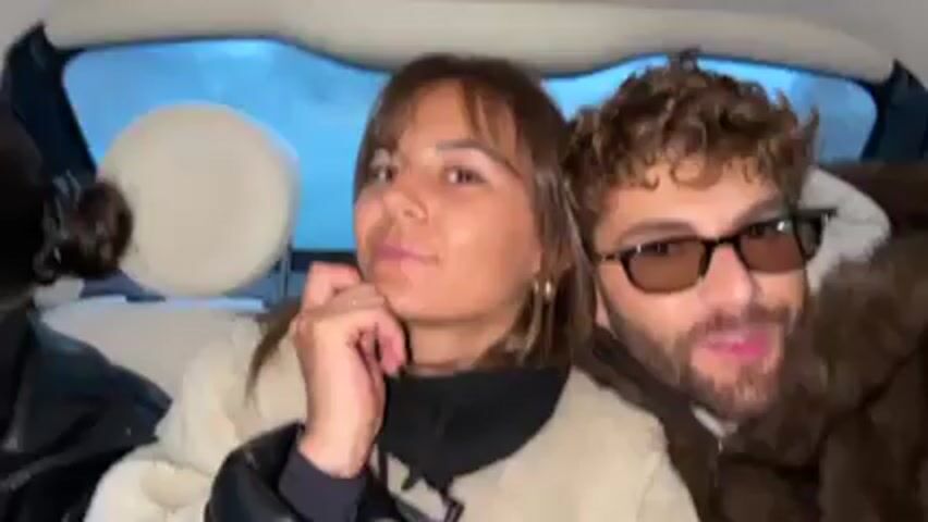A low-angle shot shows three people in the back of a car. A woman with shoulder-length brown hair sits in the center, wearing a cream-colored sherpa jacket with black leather details, a black turtleneck, and gold hoop earrings. She touches her teeth with her finger and smiles. To her right is a man with curly brown hair, a beard, and sunglasses, wearing a brown fur coat. To the woman's left, only a partial view of another person wearing a black leather jacket is visible. The car's interior is visible in the background, with light-colored leather seats. Music plays in the background.
