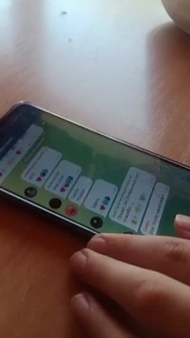 Here is a concise description of the video:

A person scrolls through messages in a Telegram application on their smartphone. The phone is lying on a light brown wooden table. The screen of the phone has some cracks. The person's left hand is visible, scrolling through the message list. Some messages in the chat are visible, including those with text and images.