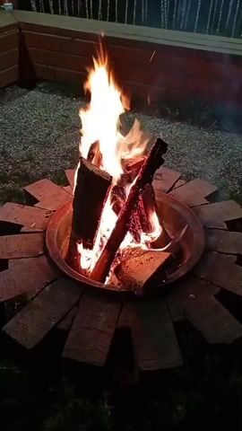 A close-up shot captures a crackling bonfire at night. The flames, a vibrant mix of oranges and yellows, dance around logs stacked in a metal fire pit. The logs are charred and burning, some with glowing embers. The fire pit is surrounded by a circular arrangement of bricks laid on a dark, gravel-like surface. In the background, out of focus, a wooden structure with lights is visible, suggesting an outdoor setting like a patio or backyard. The overall impression is one of warmth and a cozy evening ambiance.