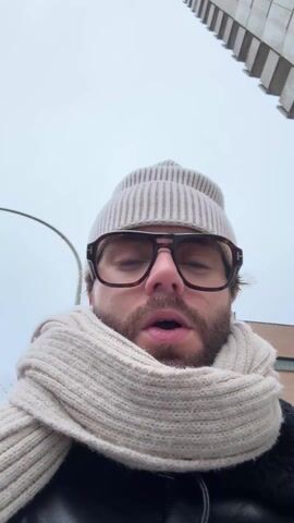 A man wearing a beige knit hat, glasses, a light-colored scarf, and a black jacket is walking outside. He also wears gloves. He adjusts his hair and looks at the camera. The background is a city street with buildings and a cloudy sky.