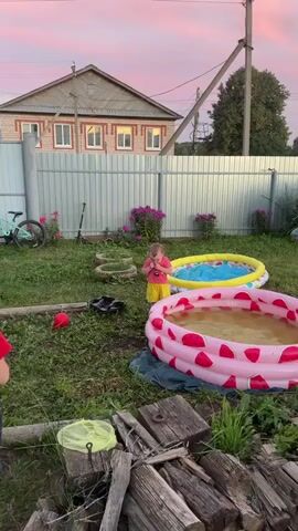 A toddler with a red t-shirt and yellow shorts stands in a grassy yard. The toddler is clapping their hands and looking to the left. In front of the toddler are two inflatable pools. The pool in the foreground is pink with a watermelon pattern and filled with murky water. Behind it, a yellow and blue inflatable pool is also visible. A white metal fence runs along the background, with a house and trees visible beyond it. The sky above is a soft pink and orange, suggesting sunset. There are purple flowers growing near the fence.