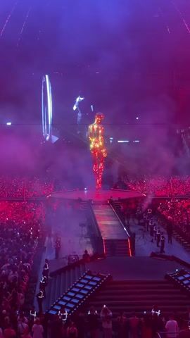 This video captures a vibrant concert scene, likely an arena performance, bathed in dramatic purple and pink lighting. At the center of the stage, a performer clad in a shimmering, gold, metallic outfit stands prominently. Behind them, a massive screen displays a large, stylized image of a person's face, also illuminated by the intense stage lighting. The audience, a dense crowd of people, fills the lower and middle sections of the venue, their presence indicated by a sea of faces and raised hands, many holding up phones to capture the moment. Smoke or fog effects add to the atmospheric ambiance, swirling around the stage and further enhancing the visual spectacle. The overall mood appears to be energetic and exciting, typical of a live musical performance. The presence of numerous uniformed individuals in black and white outfits on and around the stage suggests they are security personnel or stage crew managing the event. The visible text on a spectator's shirt in the foreground reads "81". The setting is indoors, in a large venue, and the time of day is not definitively ascertainable but is consistent with an evening concert.