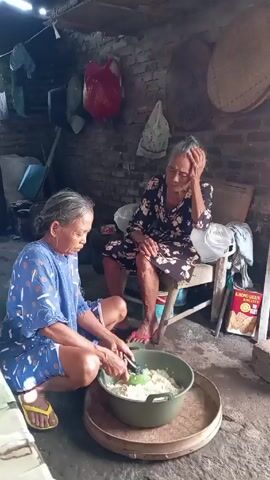 In a low-lit, modestly furnished space with brick walls, two elderly women are present. One woman, wearing a blue patterned top and shorts with yellow flip-flops, is sitting cross-legged on the floor, actively mixing a large bowl of what appears to be rice or a similar dish with a spatula. The bowl is set on a woven tray. The second woman, dressed in a dark floral dress and red flip-flops, is seated on a simple wooden chair, resting her head in her hand, appearing tired or contemplative. Behind her, there is a box of "KHONG GUAN BISCUITS," adding to the setting's domestic nature. Various household items are scattered around, including bags, containers, and woven baskets, suggesting a lived-in, functional space. The overall impression is one of quiet domestic activity.