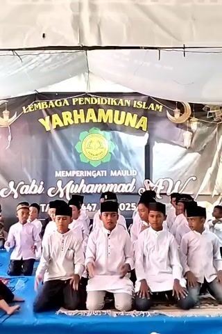 Certainly. Here is a concise description of the video:

The video shows a group of young boys, dressed in white shirts and black peci hats, seated on a blue platform indoors. They are performing a synchronized clapping and hand movement routine, likely as part of a religious or cultural event.

Behind them is a large banner that reads "LEMBAGA PENDIDIKAN ISLAM YARHAMUNA" (Islamic Education Institute Yarhamuna). The banner also commemorates the birth of the Prophet Muhammad (Nabi Muhammad SAW). 

The scene is lit by overhead lights, and the setting appears to be a temporary stage or tent structure.