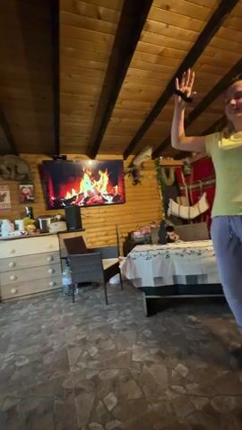 Inside a wooden-paneled room with a tiled floor, three people are dancing to music. The woman in the foreground is wearing a yellow shirt, lilac sweatpants, and maroon clogs. She is expressing herself through dance, her arms raised and mouth open. She is joined by a man in a blue shirt and dark pants and another woman in a blue shirt. They hold hands and step in unison. In the background, a TV screen displays a virtual fireplace. The room includes furniture, decorative items, and a water bottle, creating a cozy, lived-in atmosphere.