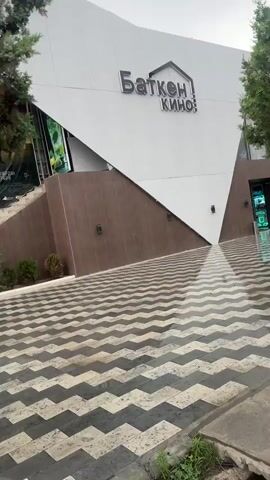 The video shows a modern cinema building with the sign "Баткен кино" (Batken Cinema) at the top. The building has white and brown sections and a distinctive geometric patterned paving stone area in front. A large movie poster is visible on the building's facade, featuring two people and a red car, with text that appears to indicate a release date of June 18th. A sign with Cyrillic text is also visible near the entrance. A tree with dark bark and green foliage stands in the foreground, partially obscuring the view of the building. The ground is wet, suggesting recent rain. The environment appears to be outdoors, with some greenery and what looks like a parking area or other outdoor space visible in the background. There are no people visible in the frames.