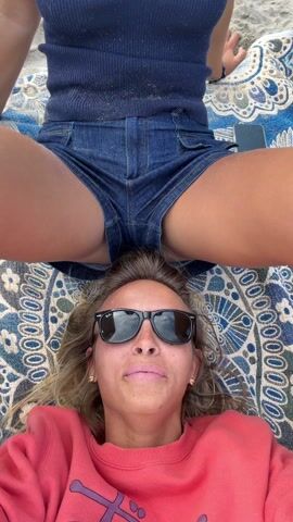 A low-angle shot shows a woman in a pink sweater lying on a blue and white patterned blanket. She is wearing black sunglasses and smiling, her white teeth visible. Another woman is sitting on the blanket behind her, facing the camera. She is wearing a dark blue sleeveless top and denim shorts. Her legs are positioned on either side of the woman lying down. A black phone is on the blanket beside her. The scene appears to be outdoors, possibly on a beach, based on the sandy texture on the legs of the seated woman.