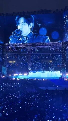 The video shows a live concert performance from a high vantage point. The stage is brightly lit with blue lights. A large screen above the stage displays a performer in close-up, while the actual performers are visible on the stage. The crowd, holding up blue lights, fills the foreground, creating a sea of blue. The backdrop of the stage includes text, and the overall atmosphere is energetic and vibrant.