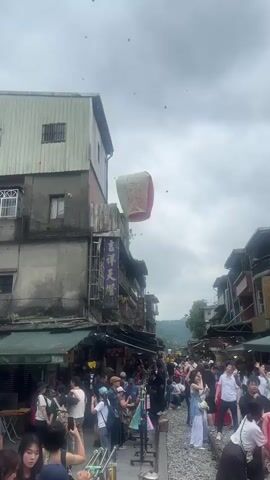 The video shows a crowded street scene, likely in Asia. People are walking along a street that appears to have train tracks running through it.  A large, square sky lantern, red, white, and green, is prominently displayed floating upwards.  Several smaller lanterns are already in the sky, scattered against a cloudy, overcast sky. Buildings are visible on either side of the street, with signs written in what appears to be Chinese characters. Many people are holding phones and taking pictures.