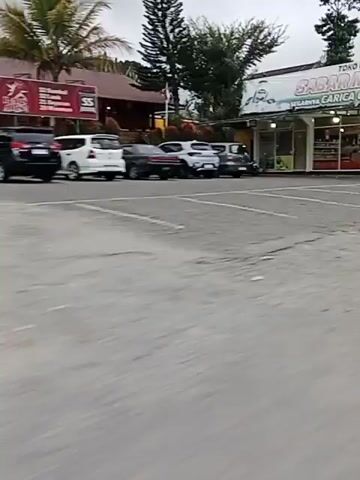 A street view footage shows several cars parked in front of buildings. One building has a sign that says "SABAR MAKMUR". Another sign reads "TOKO OLEH OLEH". There is a parking lot in front of the buildings. The video pans across the scene, showing more of the buildings and the parked cars. The environment appears to be an outdoor setting, possibly a commercial area.