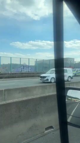 The video shows a view from inside a vehicle looking out at a highway. Several emergency vehicles are visible on the highway, including what appears to be a police car with flashing blue lights and an ambulance with flashing red and white lights. There are also civilian vehicles on the highway, including a dark-colored sedan and a red hatchback. The highway has a concrete barrier separating the lanes of traffic and a sound barrier with graffiti in the background. The sky is partly cloudy and the weather appears to be clear. The overall scene suggests an accident or other incident has occurred on the highway.