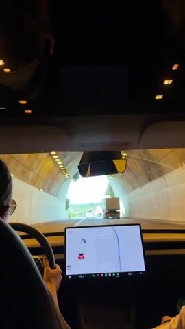 From inside a car, the video shows the driver’s perspective as the vehicle exits a tunnel. The car appears to be a Tesla, as indicated by the distinctive dashboard screen showing navigation and vehicle settings. A person’s left shoulder and part of their back are visible on the left side of the frame; their hand is lightly resting on the steering wheel. The dashboard screen is displaying a navigation map with a red car icon representing the vehicle’s position. The tunnel's interior is lit by evenly spaced lights. As the car exits, the view opens up to a highway with several other vehicles, including a large white truck. The sky is overcast, and the surrounding landscape is visible in the distance. The car’s speed is indicated as 77. The screen also shows the car is 10 km away from its destination. The interior of the car is dark, but the sunroof and front windows provide ample light to the exterior view.
