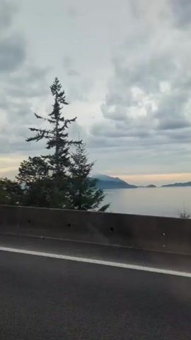 The video shows a car driving down a highway. There are tall trees on both sides of the road, and a white Jeep Grand Cherokee is driving in the right lane. The sky is overcast. The video is shot from inside the car, through the windshield. There is music playing in the background.