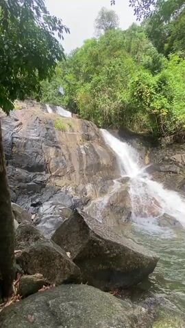 The scene features a multi-tiered waterfall cascading down a rocky hillside surrounded by lush green vegetation. Large, gray rocks are scattered at the base of the waterfall, partially submerged in the clear water of a pool. Trees with green foliage partially obscure the view on the left side of the frame. The sky is overcast, providing soft, diffused light to the scene. The sound of the flowing water is audible.