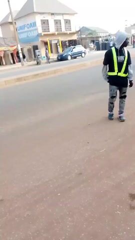 The video depicts a street scene in what appears to be a developing country, possibly in Africa. A person in a hoodie and jeans stands on a reddish, paved street. The camera pans slightly to the left, showing a street with cars, buildings, and other people in the background. The camera then focuses on two men who are shaking hands in the middle of the road. One man is wearing a dark jacket and a beanie, while the other is wearing a pink hooded sweatshirt and colorful shorts. The scene suggests a casual encounter or transaction taking place on a roadside.