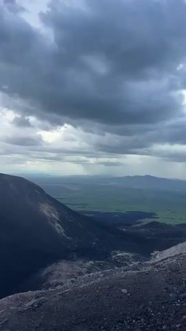 This video shows a group of people hiking up a volcanic slope. The scene transitions from a wide shot of the mountain under a cloudy sky, revealing its dark, rocky surface with patches of lighter material, to closer views of the terrain. The group is seen walking up the slope, with some individuals wearing backpacks. In the background, a vast, green landscape stretches out to the horizon under a bright, partly cloudy sky. The time of day appears to be daytime, with the lighting suggesting it could be morning or afternoon. The weather is dynamic, with dark, heavy clouds interspersed with patches of blue sky and sunlight. The text "M" is visible on the backpacks of several hikers, likely indicating a group or tour affiliation. The overall mood conveyed is one of adventure and exploration.