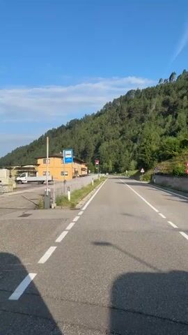 The video begins with a view of a paved road with white lane markings. On the left side of the road, there is a building with a yellow facade and some parked vehicles, including a red truck. A fence runs along the edge of the road. In the background, a steep, tree-covered hillside rises against a clear blue sky. As the video progresses, a white and blue bus passes by on the road, followed by a small, light-colored SUV. Houses are visible on the right side of the road, nestled against the hillside. Road signs, including a stop sign, are also present. The camera appears to be moving forward along the road.