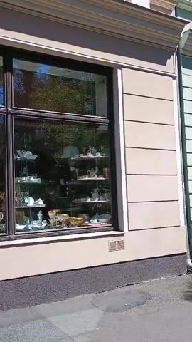 The video pans across the exterior of a store. The store has a large display window filled with glassware, chandeliers, and other decorative items. The window is divided into multiple panes by dark frames. The building exterior is light beige with horizontal detailing. A dark grey, textured foundation is visible below the window. A sidewalk with visible cracks runs in front of the store. To the right, a set of concrete stairs with brown railings leads up to a dark-colored entrance door. Above the entrance, a square sign with a yellow background and brown text reads "Porcelaine - Glas - Kristall - Bohemia." A second, smaller window is visible to the left of the entrance. Shadows are cast on the building and sidewalk, indicating a sunny day.