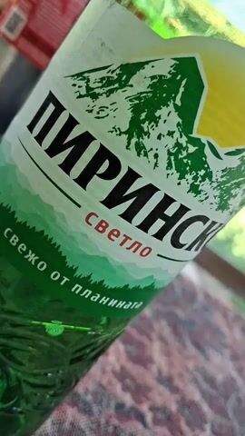The video shows a close-up of a green glass bottle with a label featuring a mountain landscape and text in Cyrillic. The label has the text "ПИРИНСКО" and "СВЕТЛО" in large black letters, with smaller text below that reads "свежо от планината". The bottle is sitting on a patterned surface, possibly a tablecloth, with other objects blurred in the background, including what appears to be another bottle and a window or outdoor scenery. The lighting suggests it's daytime.